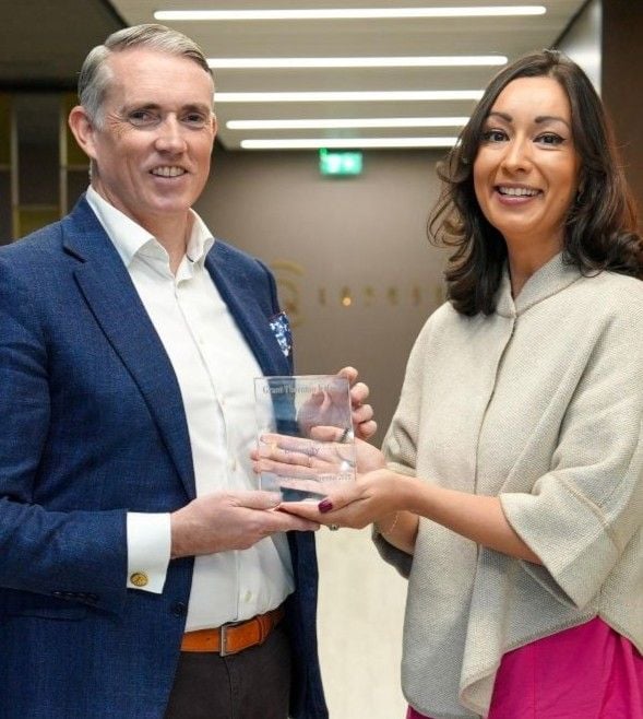 Two people standing indoors in a modern office setting, holding an award together. One person is wearing a blue blazer and white shirt, and the other is wearing a light shawl-style top with a bright pink skirt.